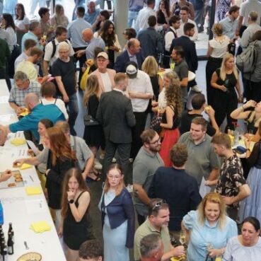 Eine große Gruppe von Menschen, die sich bei einer Veranstaltung in einem Raum mit Tischen mit Speisen und Getränken trifft. Einige Gäste essen, reden oder lachen, und die Atmosphäre wirkt lebendig und festlich.