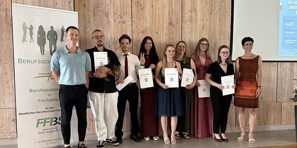 Eine Gruppe von neun Personen steht in einem Raum, lächelt und hält Urkunden in der Hand. Die meisten sind förmlich gekleidet. Sie posieren vor einer Holzwand, einem Banner und einem Projektionsschirm.