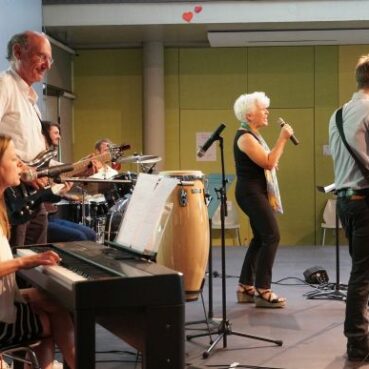 Eine Gruppe von fünf Musikern tritt in einem Innenraum auf; eine Frau spielt Keyboard, ein Mann spielt Bass, ein anderer spielt Schlagzeug, eine Frau singt mit einem Mikrofon, und ein Mann spielt E-Gitarre. Auf der Bühne sind Notenständer und Instrumente zu sehen.