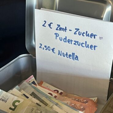 Eine offene Geldkassette mit Euro-Scheinen und -Münzen. Hinter dem Geld steht ein handgeschriebenes Schild mit den Preisen: 2€ Zimt-Zucher, Puderzucher und 2,50€ Nutella.