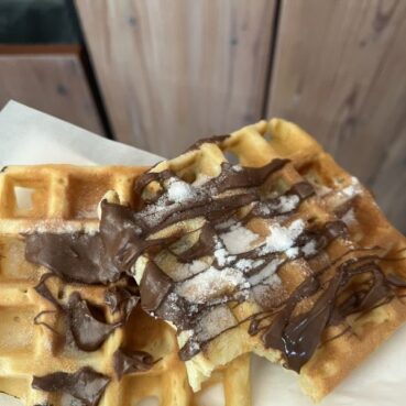 Zwei goldene Waffeln, belegt mit Schokoladensauce und Puderzucker, auf weißem Papier, mit einem hölzernen Hintergrund in der Ferne. Aus einer Waffel ist ein Stück herausgebissen worden.