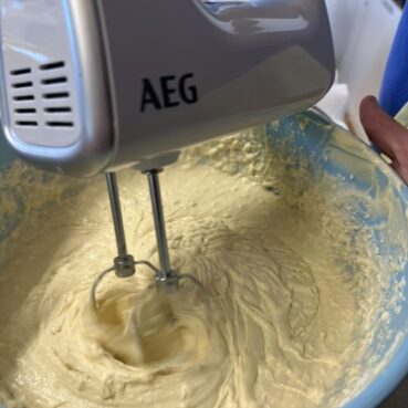 Nahaufnahme eines elektrischen Handmixers, der in einer hellblauen Schüssel einen dicken, cremigen Teig verrührt. Der Mixer ist mit AEG beschriftet und wird von einer Person in der Hand gehalten.