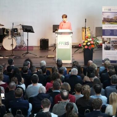 Eine Frau spricht an einem Podium mit der Aufschrift FF3S auf einer Bühne zu einem sitzenden Publikum. Auf der Bühne stehen Musikinstrumente und Ständer, und in der Nähe befindet sich eine große Anzeigetafel mit Informationen.