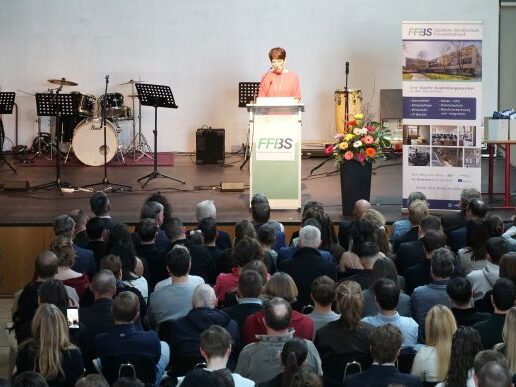 Eine Frau spricht an einem Podium mit der Aufschrift FF3S auf einer Bühne zu einem sitzenden Publikum. Auf der Bühne stehen Musikinstrumente und Ständer, und in der Nähe befindet sich eine große Anzeigetafel mit Informationen.