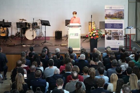 Eine Frau spricht an einem Podium mit der Aufschrift FF3S auf einer Bühne zu einem sitzenden Publikum. Auf der Bühne stehen Musikinstrumente und Ständer, und in der Nähe befindet sich eine große Anzeigetafel mit Informationen.