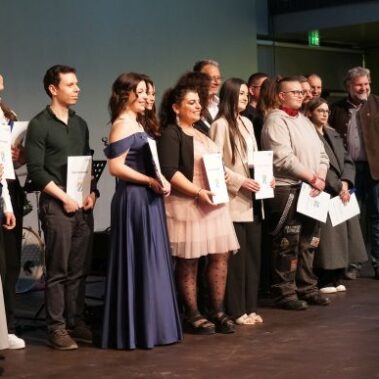 Eine Gruppe von Personen steht auf einer Bühne, einige halten Urkunden in der Hand. Sie sind formell und halbformell gekleidet, posieren und lächeln für ein Gruppenfoto vor einem blauen und grünen Hintergrund.