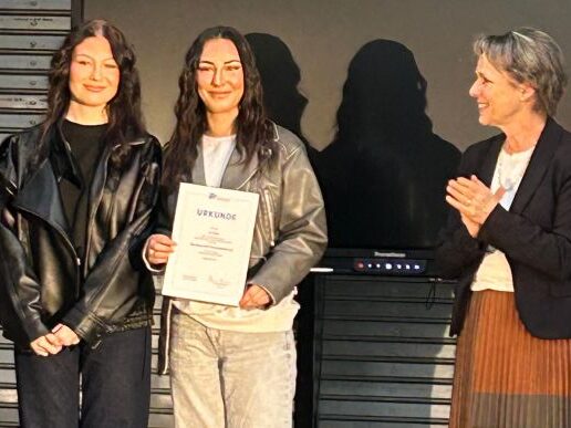 GUPP_Preisverleihung Drei Frauen stehen in einem Innenraum zusammen; die Frau in der Mitte hält eine Urkunde und lächelt, während die Frau rechts applaudiert. Alle tragen legere Jacken und sehen zufrieden aus.