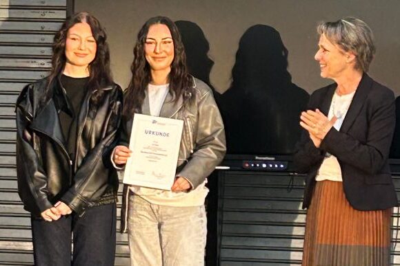 GUPP_Preisverleihung Drei Frauen stehen in einem Innenraum zusammen; die Frau in der Mitte hält eine Urkunde und lächelt, während die Frau rechts applaudiert. Alle tragen legere Jacken und sehen zufrieden aus.