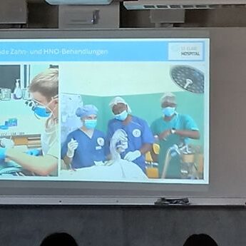 Ein Moderator steht neben einer Projektionsfläche, die Bilder von Zahnärzten bei der Behandlung von Patienten zeigt. Auf den Dias sind zwei Fotos von zahnärztlichen Eingriffen zu sehen, bei denen das medizinische Personal Masken und Handschuhe in einer klinischen Umgebung trägt.