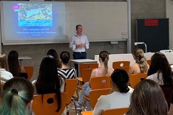 HIV-Vortrag_bearbeitet Eine Person steht vorne in einem Klassenzimmer und hält vor einer Gruppe von sitzenden Schülern einen Vortrag über HIV-Infektionen und sexuell übertragbare Krankheiten. Eine Folie mit Text und Bildern wird auf die Leinwand hinter ihr projiziert.