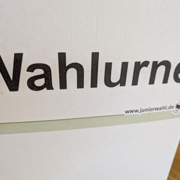 Eine Nahaufnahme einer weißen Wahlurne mit dem deutschen Wort Wahlurne in fetten schwarzen Buchstaben und der Website www.juniorwahl.de mit einem kleinen Logo darunter.