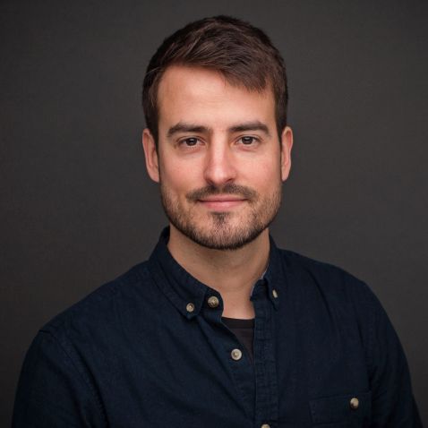 Ein Mann mit kurzen braunen Haaren, gestutztem Bart und Schnurrbart trägt ein dunkles Button-up-Hemd. Er ist der Kamera zugewandt, lächelt leicht und ist bereit, neue Kontakte zu knüpfen, während sich hinter ihm ein schlichter dunkler Hintergrund befindet.