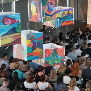 Bunt bemalte Würfel hängen von der Decke über einem sitzenden Publikum in einem großen Saal, wobei abstrakte und landschaftliche Kunstwerke auf den Würfeln zu sehen sind. Im Hintergrund befinden sich eine Bühne und eine Präsentationsleinwand.