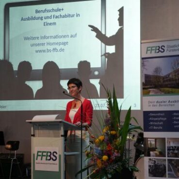 Eine Frau steht an einem Podium mit einem FFBS-Schild und spricht zu einem Publikum. Hinter ihr befindet sich ein Bildschirm mit pädagogischen Informationen und ein großes FFBS-Banner. In der Nähe sind Blumen und Musikinstrumente zu sehen.