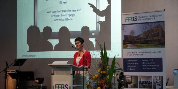 Eine Frau steht an einem Podium mit einem FFBS-Schild und spricht zu einem Publikum. Hinter ihr befindet sich ein Bildschirm mit pädagogischen Informationen und ein großes FFBS-Banner. In der Nähe sind Blumen und Musikinstrumente zu sehen.