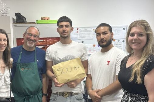 Spendenaktion_24_25 Sechs Personen stehen in einer Reihe in einem Haus und lächeln in die Kamera. Die Person in der Mitte hält einen braunen Umschlag mit der Aufschrift 500 AAH. Im Hintergrund sind Pinnwände und Schränke zu sehen.