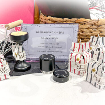 Eine Auslage mit gestreiften Geschenkschachteln, einem schwarzen Kerzenhalter, einer kleinen Dose und einem gerahmten Schild in deutscher Sprache über ein Schulprojekt für das Jahr 2025/26. Diese Gegenstände sind auf einer weißen Tischdecke angeordnet, hinter der ein Weidenkorb steht.