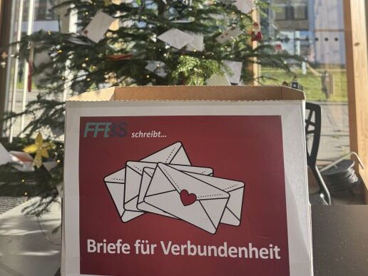 Titelbild_bearbeitet Eine Schachtel mit der Aufschrift "Briefe für Verbundenheit" und einer Illustration von drei Umschlägen steht vor einem geschmückten Weihnachtsbaum, an dem weiße Umschläge hängen. Durch die großen Fenster hinter ihnen fällt Sonnenlicht ein.