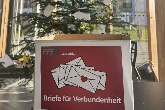 Titelbild_bearbeitet Eine Schachtel mit der Aufschrift "Briefe für Verbundenheit" und einer Illustration von drei Umschlägen steht vor einem geschmückten Weihnachtsbaum, an dem weiße Umschläge hängen. Durch die großen Fenster hinter ihnen fällt Sonnenlicht ein.