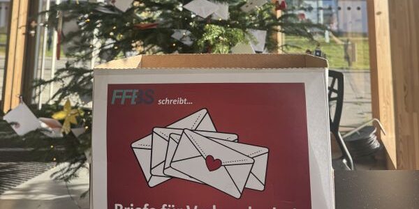 Titelbild_bearbeitet Eine Schachtel mit der Aufschrift "Briefe für Verbundenheit" und einer Illustration von drei Umschlägen steht vor einem geschmückten Weihnachtsbaum, an dem weiße Umschläge hängen. Durch die großen Fenster hinter ihnen fällt Sonnenlicht ein.