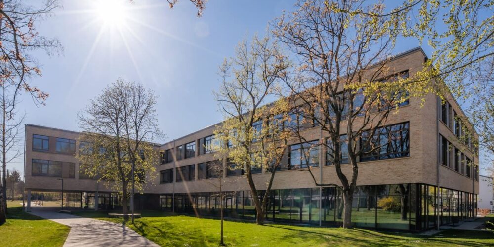 Das moderne, dreistöckige Schulgebäude der Berufsschule Fürstenfeldbruck mit großen Fenstern steht unter strahlendem Sonnenschein und blauem Himmel, umgeben von grünem Gras, Bäumen und einem gepflasterten Weg.