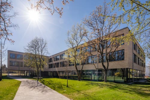 berufsschule-fuerstenfeldbruck-schulgebaude-500×333 Das moderne, dreistöckige Schulgebäude der Berufsschule Fürstenfeldbruck mit großen Fenstern steht unter strahlendem Sonnenschein und blauem Himmel, umgeben von grünem Gras, Bäumen und einem gepflasterten Weg.