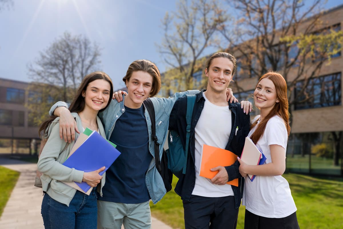 Vier lächelnde Schüler stehen im Freien auf dem Campusweg der Berufsschule Fürstenfeldbruck, in der Hand Mappen und Hefte. Zwei haben ihre Arme um die Schultern des anderen gelegt. Im Hintergrund sind Bäume und ein modernes Backsteingebäude zu sehen.