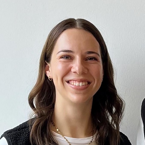 Eine junge Frau mit langen braunen Haaren lächelt in die Kamera. Sie trägt ein weißes Oberteil und eine dunkle Weste, dazu eine einfache Halskette, und steht vor einem schlichten weißen Hintergrund.