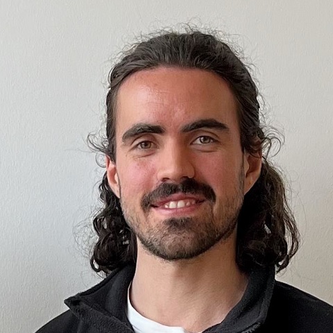 Ein Mann mit langen lockigen braunen Haaren, einem Schnurrbart und einem Bart lächelt in die Kamera. Er trägt eine schwarze Jacke über einem weißen Hemd und steht vor einer schlichten hellen Wand.