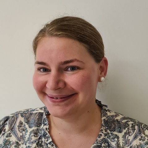Lächelnde Frau mit heller Haut und zurückgebundenem blondem Haar, die eine gemusterte Bluse und Perlenohrringe trägt, steht vor einem schlichten weißen Hintergrund.